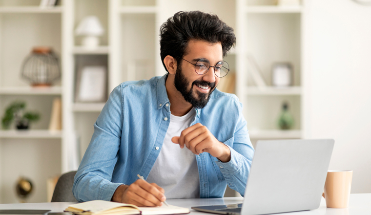 Online Education. Young Indian Man Watching Webinar On Laptop Computer And Taking Notes, Smiling Eastern Millennial Guy Enjoying Distance Learning, Sitting At Desk At Home Office, Free Space