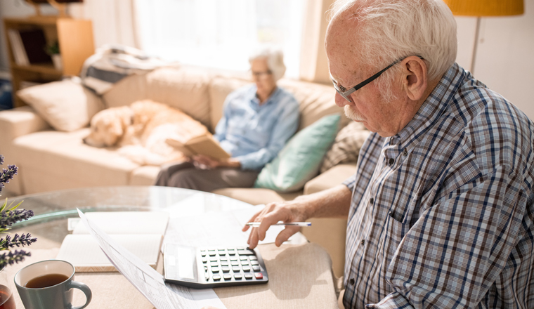 Side view portrait of senior man filling forms and paying taxes while sitting at table with elderly woman and pet dog sitting on couch in background, copy space