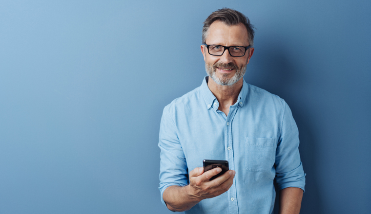 Smiling friendly man wearing glasses standing holding his mobile phone looking at the camera with a warm smile over a blue studio background with copy space