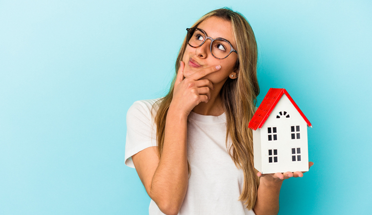 Young caucasian woman holding a house model isolated on blue background looking sideways with doubtful and skeptical expression.