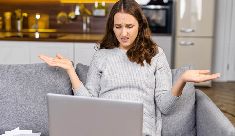 Confused female entrepreneur sits on the couch and staring at the laptop screen, young woman shrugs shoulders, does not understand what happened, system error on the computer