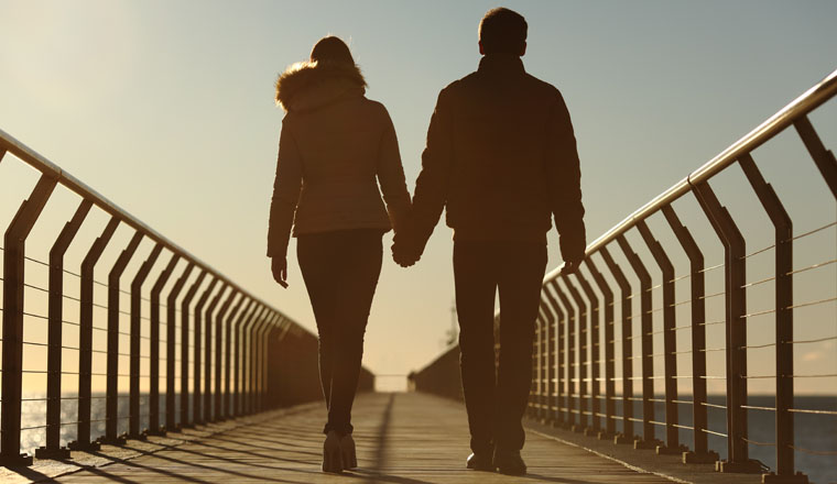 Back silhouette of a couple walking holding hands on a bridge in the beach at sunset