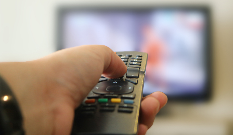 Image of a hand clicking TV Remote Control with out of focus television in the background