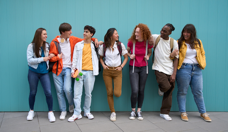 Multiracial group of cheerful erasmus students enjoying free time. Young friends together leaning against a blue wall and looking at each other smiling. Generation z and youth relationships.