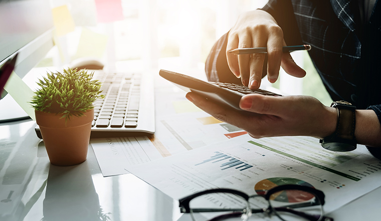 Close up of businessman or accountant hand holding pen working on calculator to calculate business data, accountancy document and laptop computer at office, business concept