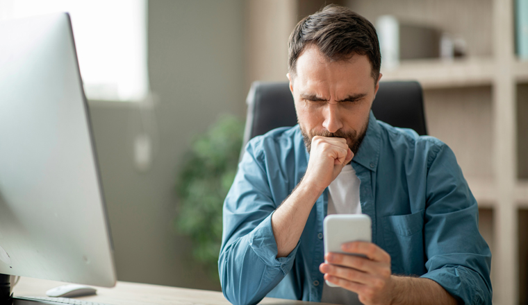Stressed businessman looking at smartphone screen while sitting at desk in office, worried male entrepreneur reading sms and frowning, received bad news, got scam message, closeup shot
