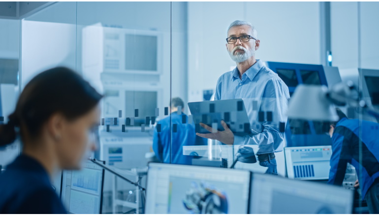 Modern Factory: Senior Engineer Looks at Camera, Holding Laptop, Inspecting, Overlooking Workshop Operation. Manufactory Has Professionals Working on CNC Machinery, Robot Arm.