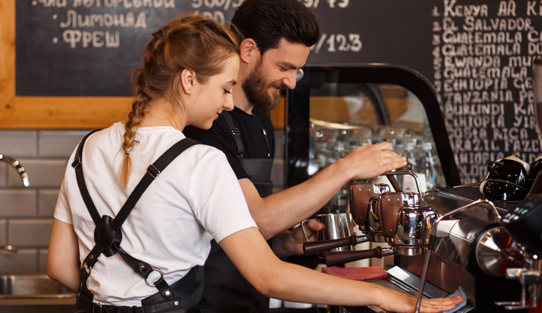 Two young smiling barista at work. Professional barista team brewing coffee using coffee machine in coffee shop. Happy young man and woman developing own coffee business. Coffee shop concept.