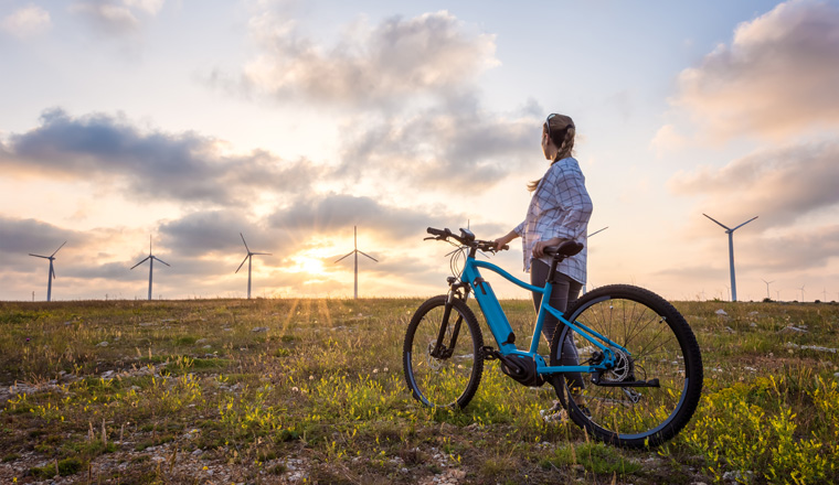 A woman with a bike enjoys the view of sunset over a summer field with a wind farm