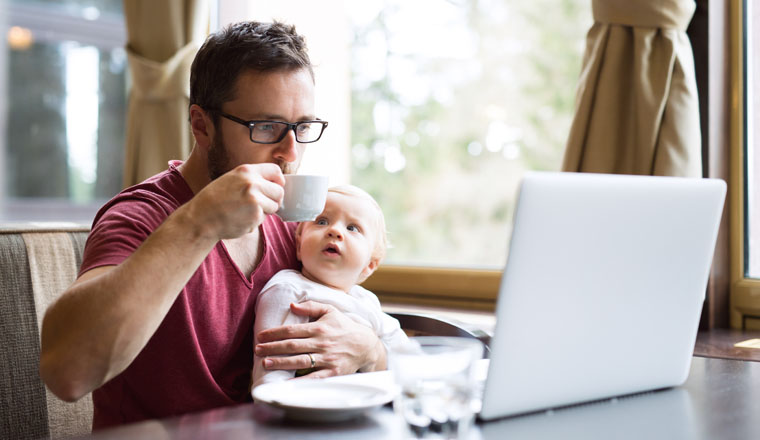 Young handsome man with notebook in cafe sitting at the table drinking coffee, holding his son in his lap