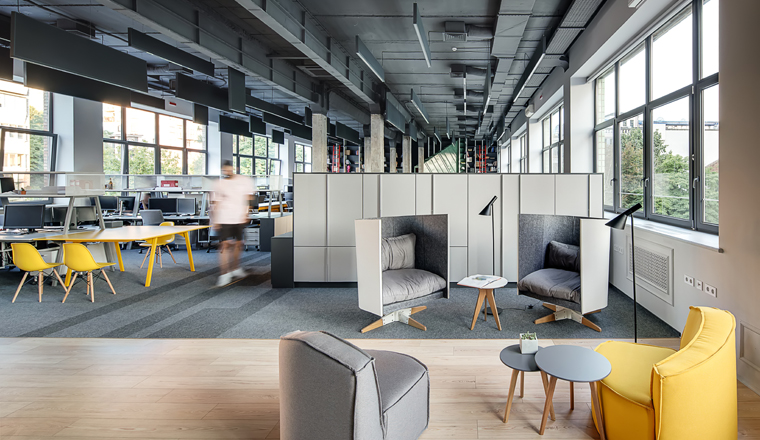 Office in a loft style with large windows, gray walls and concrete columns. There are many workplaces with computers and shelves and lockers, relax zone with armchairs with round tables and lamps.