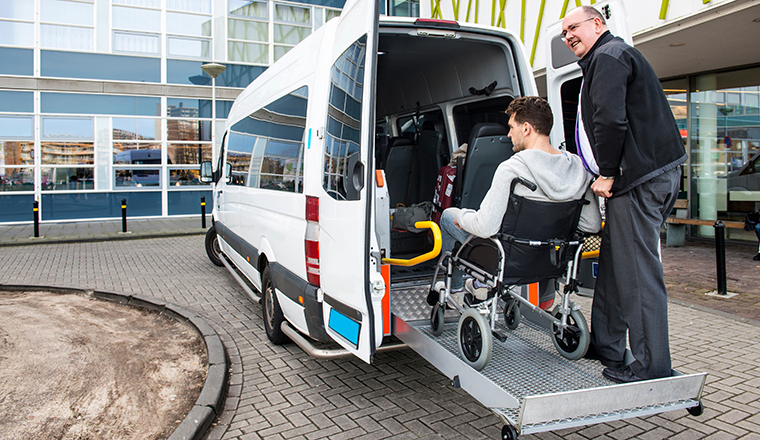 The driver of a wheel chair taxi, helping a disabled man in a wheel chair, using the lift in the back of his mini van