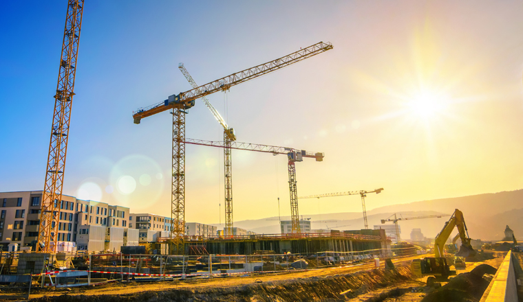 Large construction site including several cranes working on a building complex, with clear blue sky and the sun