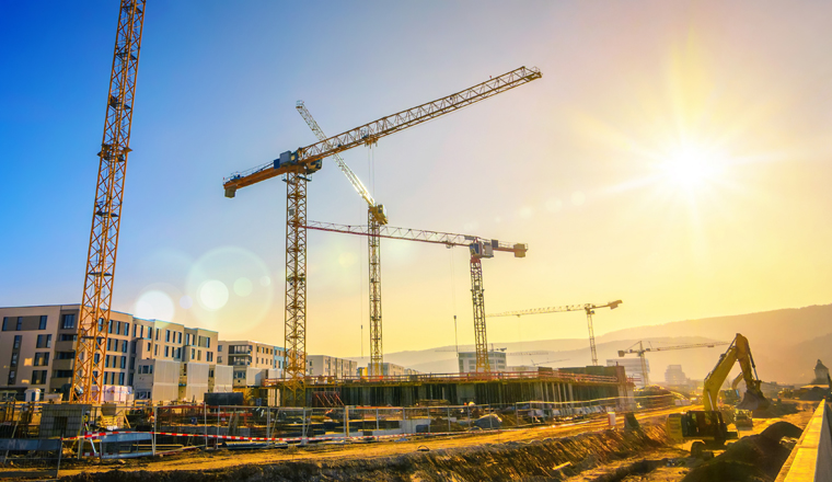 Large construction site including several cranes working on a building complex, with clear blue sky and the sun