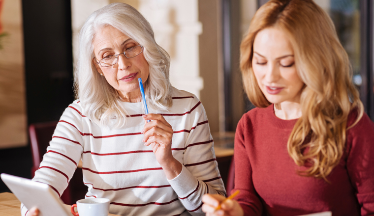 Smart women. Clever attentive senior woman sitting at the table and touching her lips with a pen while looking at the screen of a modern tablet with a young responsible designer sitting by her side