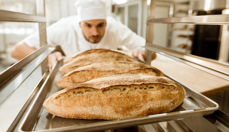 young baker putting trays of fresh bread on stand at baking manufacture