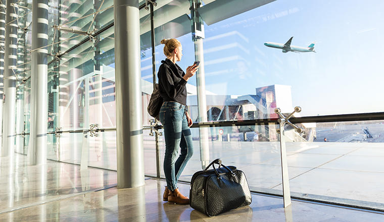 Young casual female traveler at airport, holding smart phone device, looking through the airport gate windows at planes on airport runway.