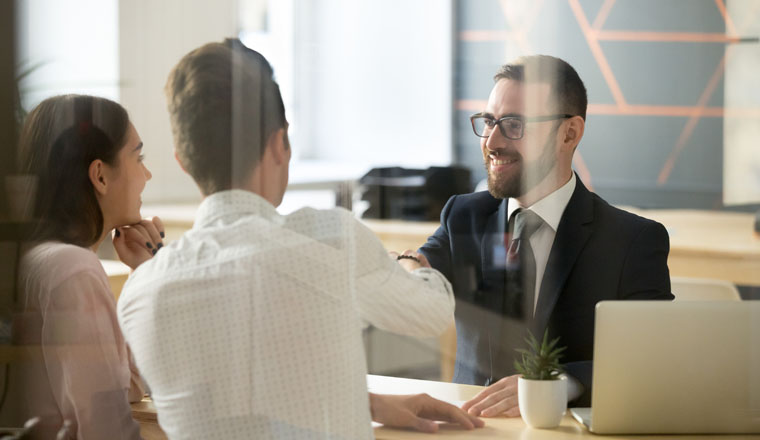 Smiling male realtor or broker shaking hand of excited buyers couple, negotiating about first house purchase or taking loan, insurance agent welcoming clients with handshake at consultation meeting