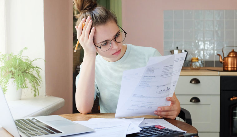 Indoor shot of young European Caucasian girl looking at financial documents at home with deeply bored face looking sick and tired of her economic problems, trying to check counts and all details