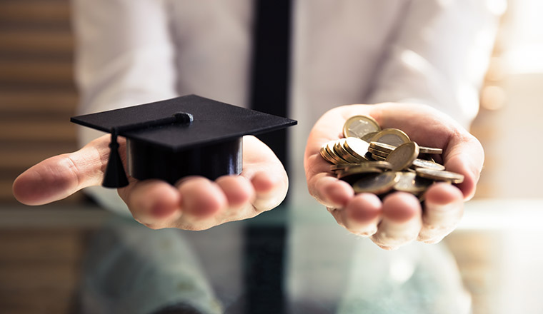 Human Hand Holding Black Graduation Hat And Golden Coins Over Desk