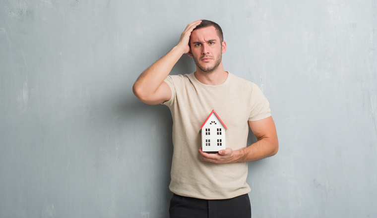 Young caucasian real state agent man over grey grunge wall holding house stressed with hand on head, shocked with shame and surprise face, angry and frustrated. Fear and upset for mistake.