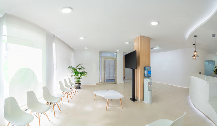 Panorama of a bright reception and waiting room in a clinic with desk, modern chairs and plants. Indoor mockup with screen with copy space.