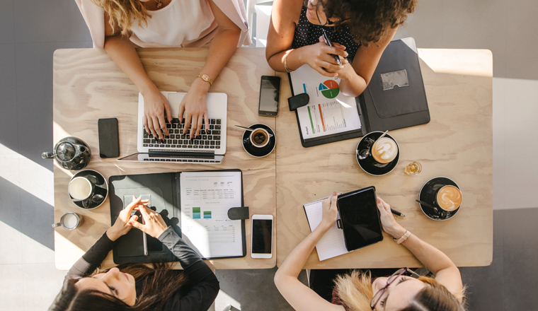 Top view of four women sitting around table in cafe with laptop, digital tablet and documents. Group of women working together in coffee shop.