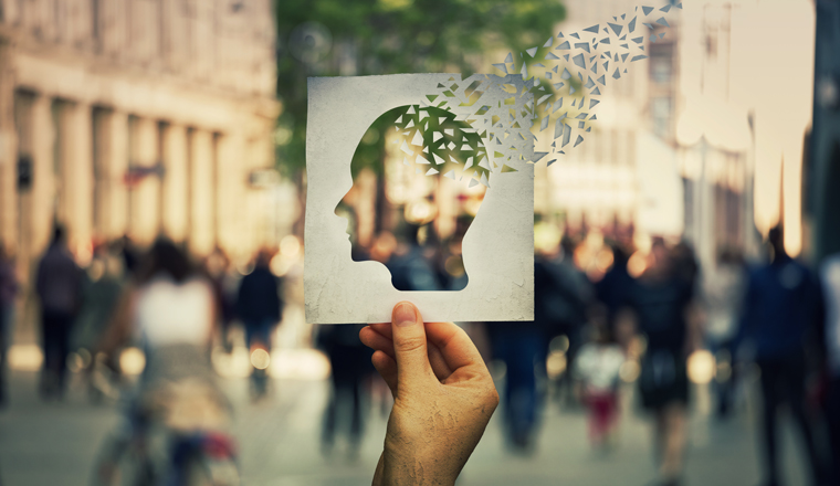 Hand holding a paper sheet with human head icon broken into pieces over a crowded street background. Concept of memory loss and dementia disease. Alzheimer's losing brain and memory function.