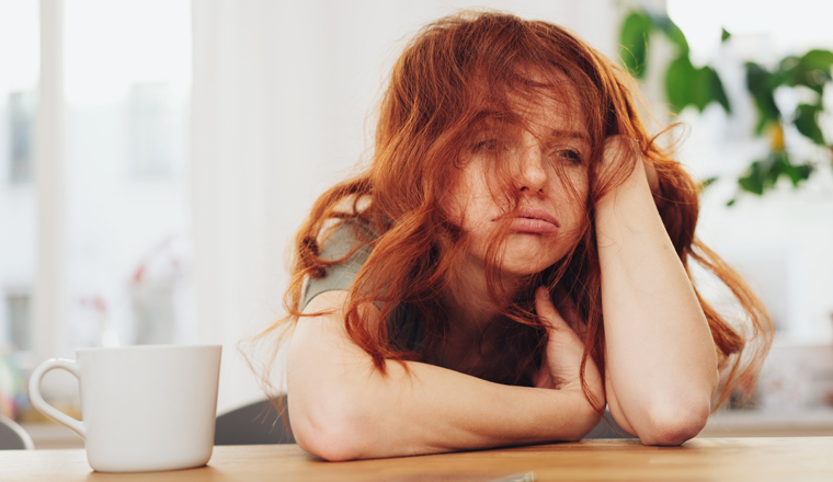 Red-haired girl sitting at the table indoors with boring face and looking away through messy hair, leaning on her elbow