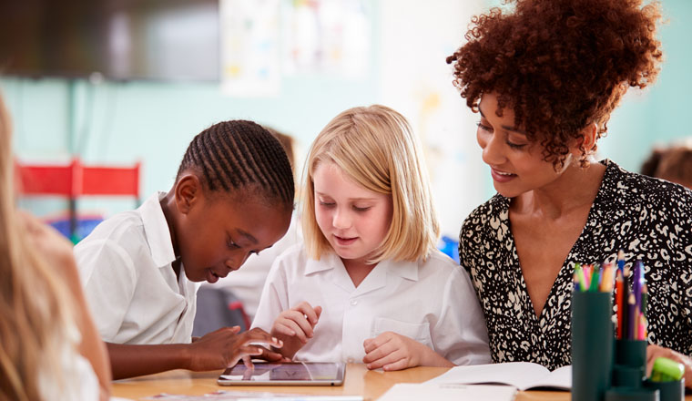 Female Teacher With Two Elementary School Pupils Wearing Uniform Using Digital Tablet At Desk