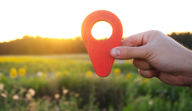 A hand is holding a red location marker in the sunset background. The concept of tourism and travel. Navigation and exploration. Destination. Holiday, vacation. Buying building land. Selective focus