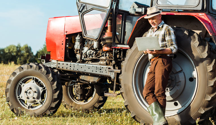 panoramic shot of farmer in straw hat using laptop near tractor