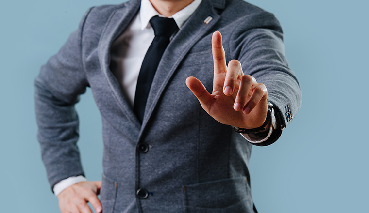 Businessman in a gray suit is making objection gesture by extending hand with the index finger up, holding his second hand on the waist over blue background. Cropped, no head.