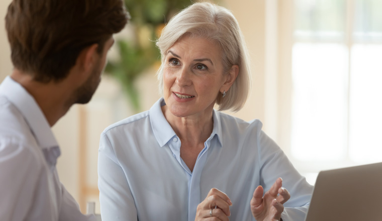 Middle aged old businesswoman manager mentor speaking to businessman client offering insurance banking services deal teaching intern or convincing customer at business office meeting sit at table.