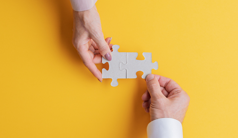 Wide view image of male and female hand joining two matching puzzle pieces together in a conceptual image of teamwork and cooperation. Top view with copy space.