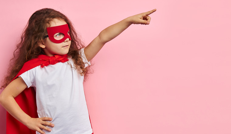 Attractive cute kid wearing red superhero suit and mask stand showing side isolated ove pink background