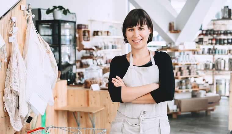 Portrait of owner of sustainable small local business. Female seller assistant of zero waste shop on interior background of shop. Smiling young woman in apron standing in plastic free grocery store.