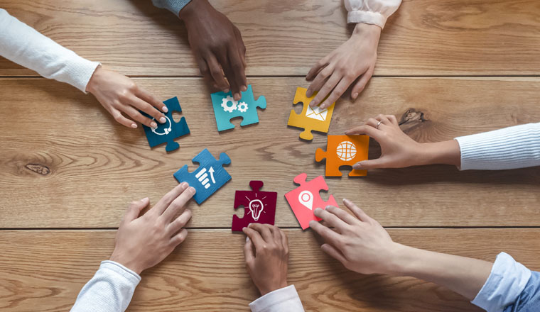 Hands of international coworkers sitting around table, putting colorful puzzles together, teamwork concept, top view