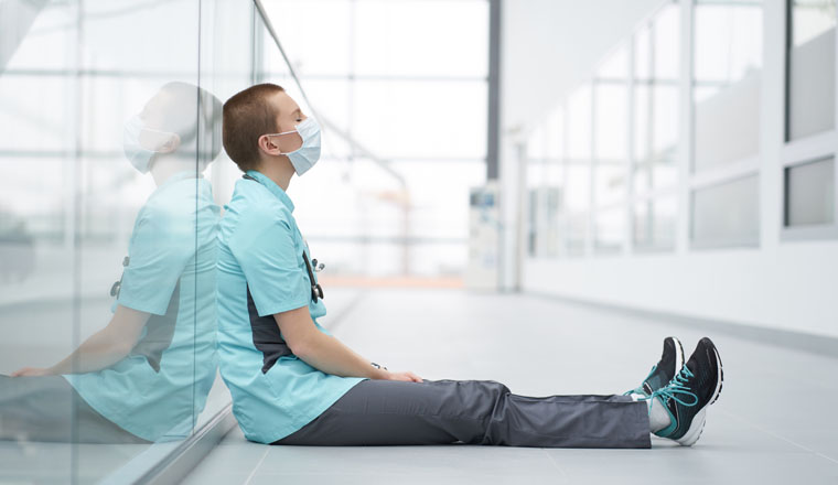Burnout concept. Exhausted caucasian female short haircut doctor, wearing medical mask, sitting on a floor in corridor of clinic with closed eyes