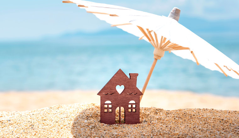 Miniature house and umbrella on beach, blue sea and sky on blurred background. Vacation home for happy holiday for family. Real estate, sale or property investment concept. Closeup. Copy space