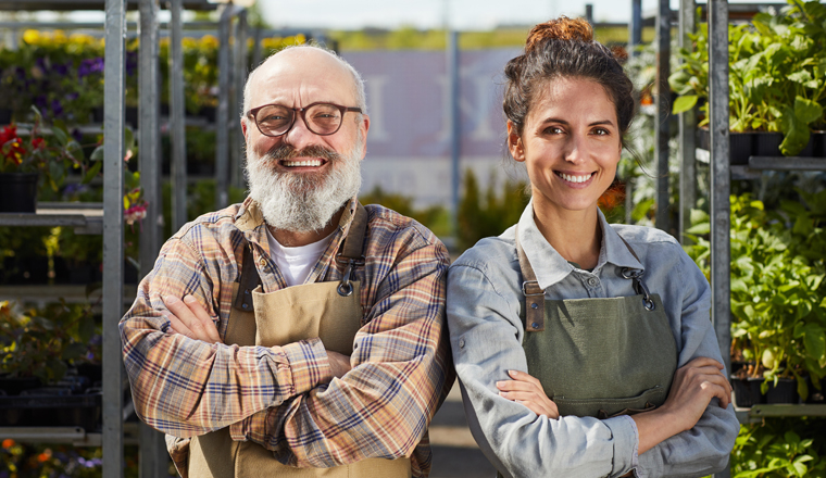 Waist up portrait of smiling bearded farmer with young female worker looking at camera and smiling happily while standing in greenhouse at plantation lit by sunlight