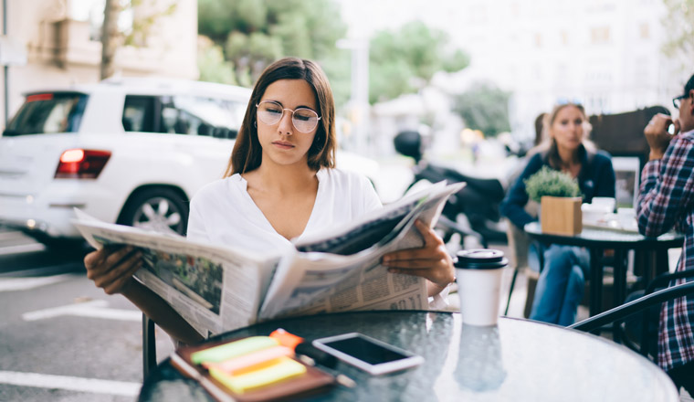 Serious young female in eyeglasses and white shirt sitting in city street at small table outside cafe and looking through newspaper