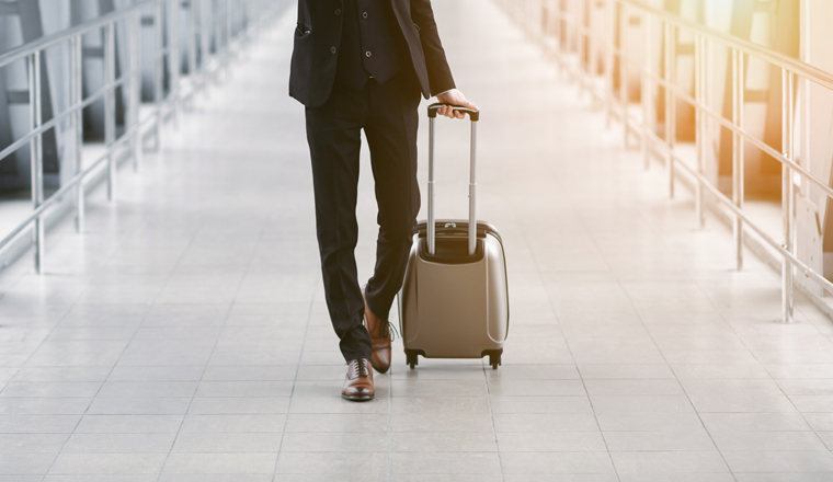 Business Trip. Frontal view of unrecognizable guy in suit pulling trolley bag walking in the airport, copy space, sun flare