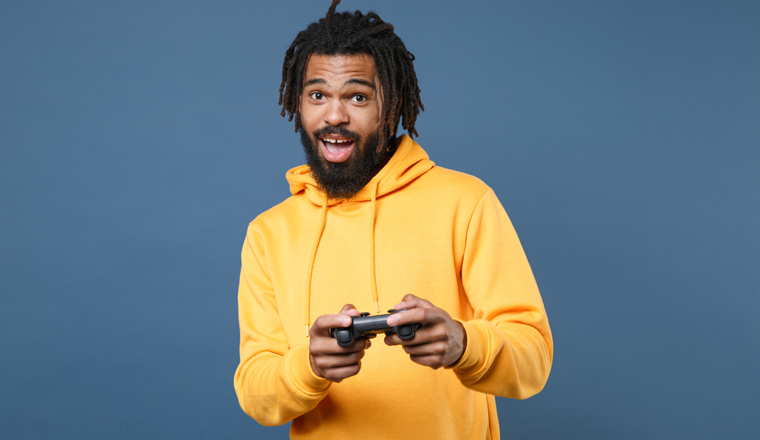 Excited young african american man guy in casual yellow streetwear hoodie isolated on blue wall background studio portrait. People sincere emotions lifestyle concept. Play game with joystick