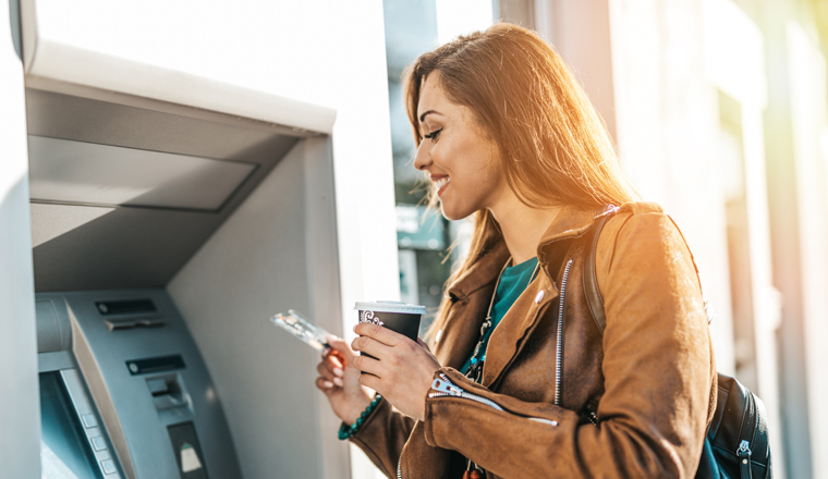 Happy young adult woman standing in front of ATM machine, smiling and holding credit or debit card. 