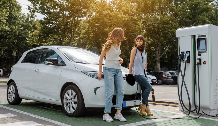 Two happy young beautiful women are talking to each other until their electric car is charging at the charging station situated in the car park. Eco friendly transport concept