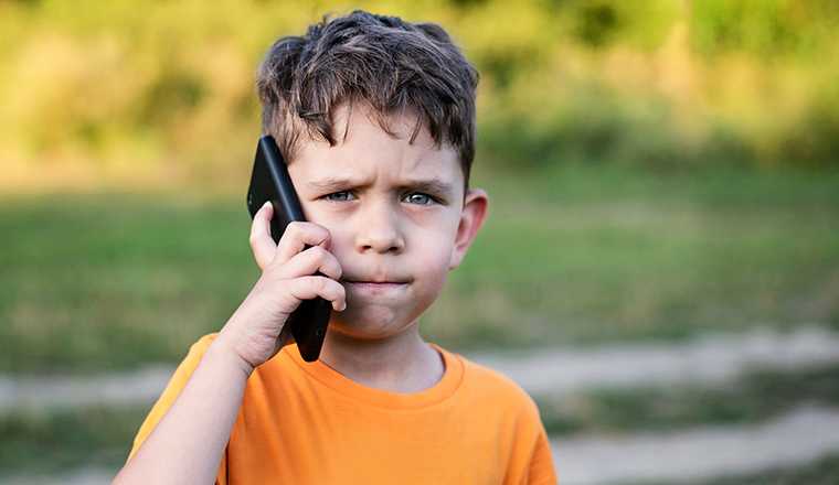The little boy is talking on the phone with a serious face in a park, trying to explain to his dad that he misses him very much, his father had to go to another country to earn money