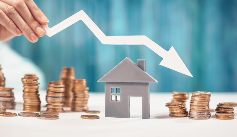 Woman holds an arrow down over model of the house and stack of coins. Concept of the crisis in the real estate market.