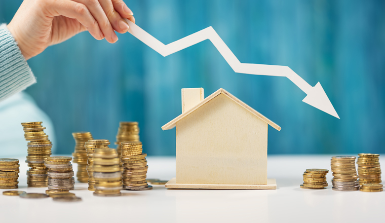 Woman holds an arrow down over model of the house and stack of coins. Concept of the crisis in the real estate market.