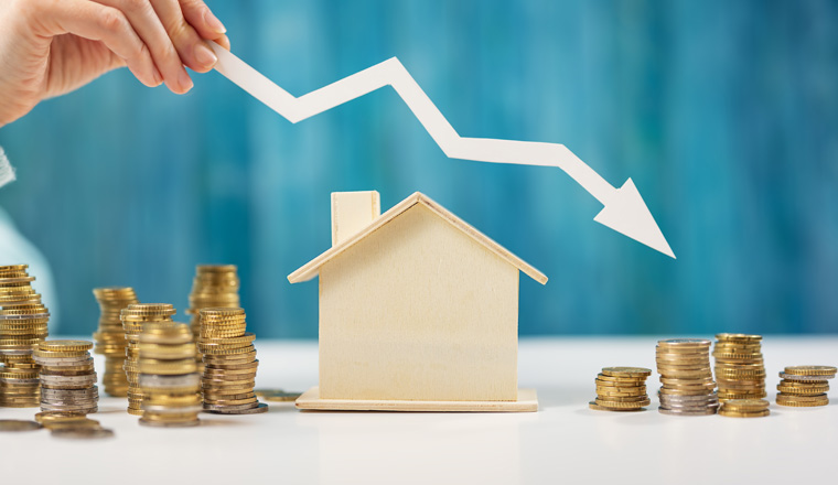 Woman holds an arrow down over model of the house and stack of coins. Concept of the crisis in the real estate market.